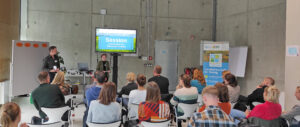 Wissenschaftler und Landwirte in einem Raum. Sie sind von hinten zu sehen. Sie sprechen über Wissenstransfer zwischen Forschung und Praxis. Gegenüber steht ein Bildschirm, der das Thema der Diskussionsrunde zeigt. Eine Frau ist von vorne zu sehen, sie spricht zu den Menschen.