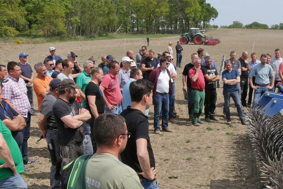 40 Landwirte stehen auf einem Acker und blicken auf ein landwirtschaftliches Gerät. Sie sprechen über das Gerät, eine Rollhacke, und tauschen Für und Wider aus.