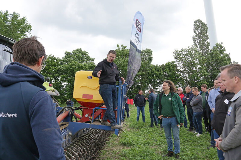 Zahlreiche Landwirte haben sich um einen Traktor, der mit Rollhacken bestückt ist, versammelt. Oben auf dem Anbbaugerät steht der Verkäufer der Technik und diskutiert mit den Umstehenden. Foto: Julian Delbrügge (ILU)
